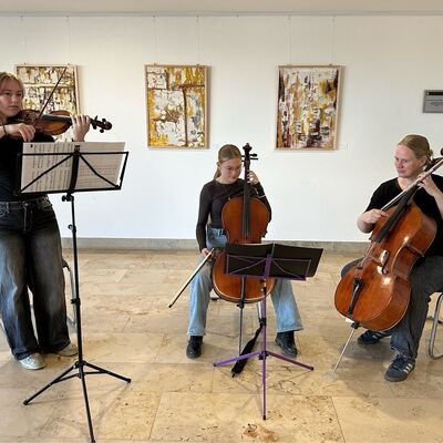 Zwei M&auml;dchen im Teenageralter und eine Frau musizieren gemeinsam. Im Hintergrund h&auml;ngen an der Wand hochformatige Bilder mit Naturmaterialien gefertigt.