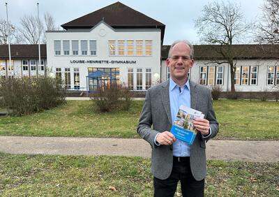 Bildungsdezernent Holger Mittelstädt hält die neue Schulbroschüre in der Hand. Im Hintergrund steht das Louise-Henriette-Gymnasium Oranienburg.