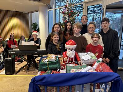 Ein Wagen voller Geschenke, glückliche Kinder und im Hintergrund ein Weihnachtsbaum sowie der Kinderchor der Kreismusikschule.