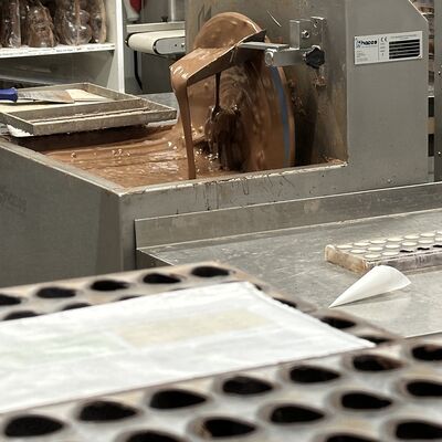 Im Bildfokus ist geschmolzene Schokolade in einem Beh&auml;lter. Vordergr&uuml;ndig sind unscharf mit Schokolade gef&uuml;llte Pralinenf&ouml;rmchen zu sehen.
