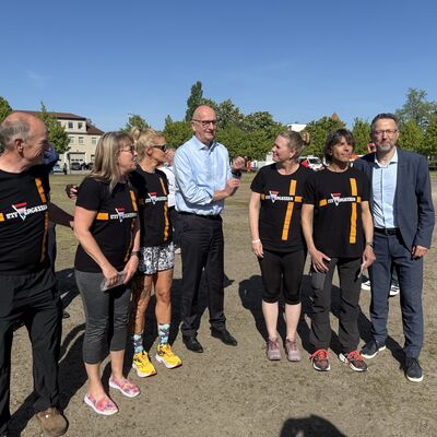 Landrat Alexander T&ouml;nnies, mit grauem Sakko und grauer Hose, steht zusammen mit dem brandenburgischen Ministerpr&auml;sidenten Dietmar Woidke, der ein hellblaues Hemd und eine schwarze Anzughose tr&auml;gt, und f&uuml;nf L&auml;uferinnen und L&auml;ufern auf einem Sandboden beim Sachsenhausener Gedenklauf 2025.