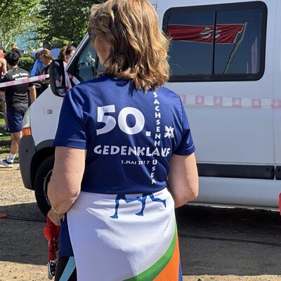 Im Vordergrund steht eine weibliche Zuschauerin mit dem R&uuml;cken zur Kamera. Sie tr&auml;gt ein blaues T-Shirt mit der Aufschrift "50. Sachsenhausener Gedenklauf". Im Hintergrund steht ein wei&szlig;er Transporter.