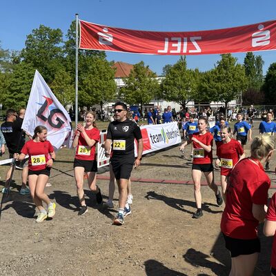 Vier M&auml;dchen mit kurzen roten T-Shirts und schwarzen Shorts &uuml;berqueren die Ziellinie beim Sachsenhausener Gedenklauf. In der Mitte ist ein m&auml;nnlicher L&auml;ufer mit kurzem, schwarzem Shirt und kurzer, schwarzer Hose sowie dunkler Sonnenbrille zu sehen.
