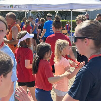 Im Vordergrund sind mehrere junge M&auml;dchen des Sachsenhausener Gedenklaufs mit kurzen T-Shirts und kurzen Hosen zu sehen, die laufen am Stand des Kreissportbundes vorbeilaufen.