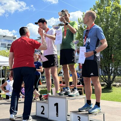Auf einem Podium stehen drei M&auml;nner, die jeweils einen Pokal und eine Urkunde in den H&auml;nden halten und eine Siegermedaille um den hals tragen. Auf dem Podium steht in der Mitte die Zahl 1, rechts die Zahl 3 und links die Ziffer 2.