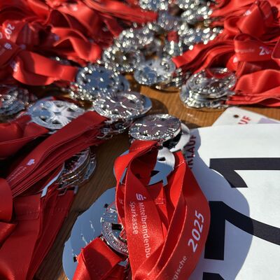 Auf einem Tisch liegen zahlreiche, silberfarbene Teilnehmermedaillen des Sachsenhausener Gedenklaufs mit rotem Schl&uuml;sselband mit dem Aufdruck "Mittelbrandenburgische Sparkasse 2025".