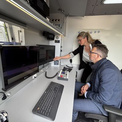 Ein Mann mit einer Brille im blauen Anzug sitzt an einem Schreibtisch mit Tastatur und zwei Monitoren. Neben steht eine blonde Frau mit Brille mit silberfarbener Bluse und schwarzem Jacket, die mit der rechten Hand auf einen der beiden Monitore zeigt.