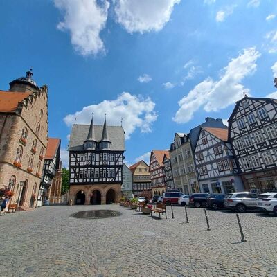 Das Bild zeigt einen Platz mit Fachwerkh&auml;usern und gepflastertem Boden unter blauem Himmel mit vereinzelten Wolken.
