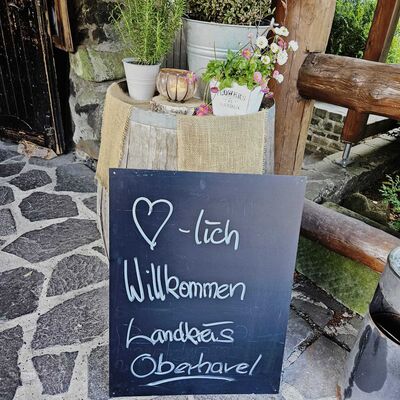 Ein gestaltetes Willkommensschild mit der Aufschrift &raquo;Herzlich willkommen Landkreis Oberhavel&laquo; steht vor einem Holzfass, dekoriert mit Blumen und Kr&auml;utern.