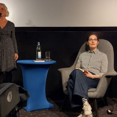 Eine Frau mit kinnlangen, blonden Haaren steht mit einem Mikrofon in der rechten Hand links auf dem Bild neben einem blauen Tisch. Rechts im Bild sitzt eine Frau mit Brille und braunen Haaren in einem hellgrauen Sessel und hat ein Buch auf dem Scho&szlig;.