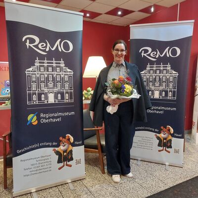 Eine Frau mit Brille und dunkelbraunen, zum Zopf gebundenen Haaren steht mit einem Blumenstrau&szlig; in den H&auml;nden vor zwei Roll-up Bannern des ReMO - Regionalmuseum Oberhavel. Bei der Frau handelt es sich um die Autorin Alex Beer.