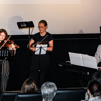 Drei Sch&uuml;lerinnen der Kreismusikschule spielen auf Geigen und einem Keyboard vor dem Publikum. Im Hintergrund befindet sich eine beleuchtete Leinwand.