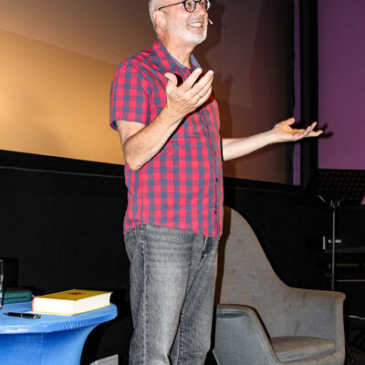 Peter Grandl steht vor dem Publikum zwischen Sessel und dem Beistelltisch. Er tr&auml;gt ein schwarz-rot kariertes Hemd und eine graue Jeans.