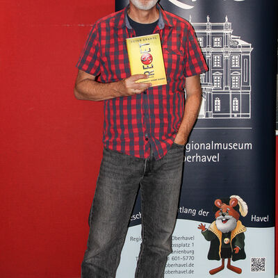 Der Autor Peter Grandl vor dem Roll-up Banner des ReMO - Regionalmuseum Oberhavel nach seiner Lesung am 30.06.2025 w&auml;hrend er sein Buch "Reset" hochh&auml;lt.