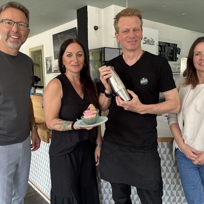 Zwei M&auml;nner und zwei Frauen stehen vor einem Caf&eacute;tresen und l&auml;cheln in die Kamera.