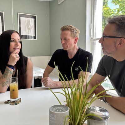 Eine Frau und zwei M&auml;nner sitzen an einem wei&szlig;en Caf&eacute;tisch und unterhalten sich. Auf dem Tisch steht ein Longdrinkglas mit einem gelben Getr&auml;nk.