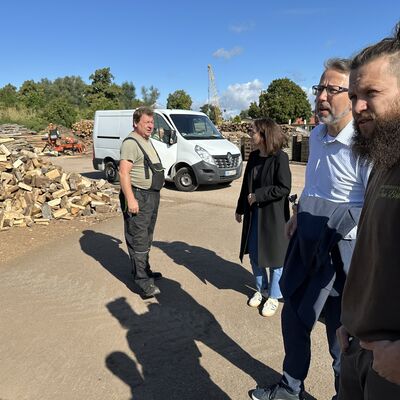 Im Vordergrund rechts im Bildrand zwei M&auml;nner, die seitlich an der Kamera vorbeischauen. Im Hintergrund rechts ein &auml;lterer Mann vor einem Haufen gespaltenem Kaminholz.