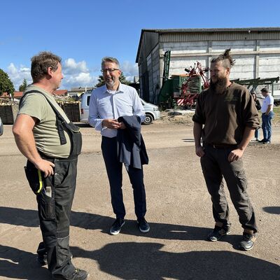 Drei M&auml;nner stehen zusammen und unterhalten sich. Der Mann in der Mitte tr&auml;gt ein blaues Hemd, eine Brille und eine dunkle Stoffhose. Die anderen beiden M&auml;nner tragen lockere, landwirtschaftliche Arbeitskleidung.