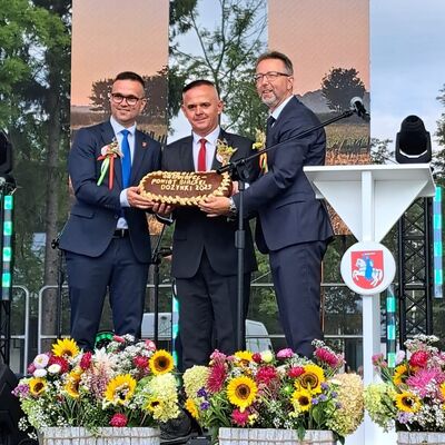 Drei M&auml;nner in Anz&uuml;gen und mit Krawatte stehen auf einer B&uuml;hne und halten ein Brot in die Kamera.