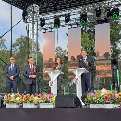 Drei M&auml;nner und eine Frau stehen auf einer B&uuml;hne mit Blumengestecken im Vordergrund und vier rechteckigen Deckenbannern im Hintergrund.