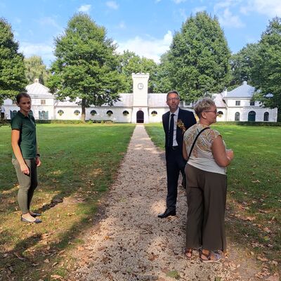 Zwei Frauen und ein Mann stehen auf einem Weg. Links und rechts Wiese und B&auml;ume. Im Hintergrund ein Gutsschloss und Pferdegest&uuml;t.