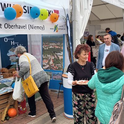 M&auml;nner und Frauen stehen im Bildvordergrund. Eine Frau mit einer gelben Umh&auml;ngetasche beugt sich &uuml;ber einen mit Brot gedeckten und bunten Luftballons geschm&uuml;ckten Stand. Unterhalb der Luftballons h&auml;ngt ein auf polnisch bedrucktes Banner auf dem steht: Powiat Oberhavel (Niemcy).