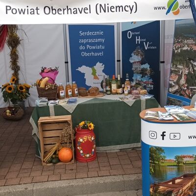 Frontaler Blick auf einen herbstlich dekorierten Messestand an dem es Sonnenblumen, K&uuml;rbisse, Gem&uuml;se, S&auml;fte, Brot gibt. Im Vordergrund steht ein Stehpult mit Flyern, das mit einem Bildmotiv und Logo des Landkreises Oberhavel bedruckt ist.