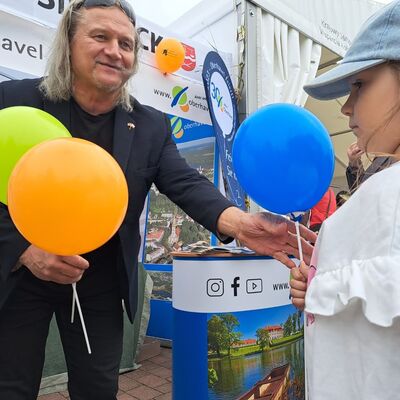 Ein Mann in schwarzem Shirt und mit schwarzem Sakko h&auml;lt in der rechten Hand einen orangefarbenen und einen hellgr&uuml;nen Luftballon.  Mit links &uuml;berreicht er einem M&auml;dchen mit einer lang&auml;rmeligen, wei&szlig;en Bluse und einem hellblauen Basecap einen dunkelblauen Luftballon.