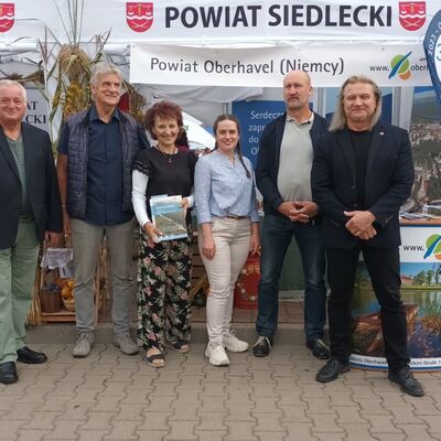 Eine Gruppe von vier M&auml;nnern und zwei Frauen steht vor einem Messestand, auf dem mit polnischer Schrift Powiat Siedlecki und Powiat Oberhavel (Niemcy) steht. Die Personen schauen frontal in die Kamera.