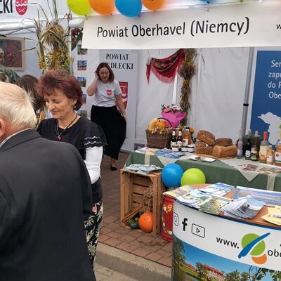 M&auml;nner und Frauen stehen links im Bildvordergrund. Eine Frau mit einem schwarzem Rock im Bildhintergrund steht neben dem gedeckten Tisch des Messestandes, auf dem es typische K&ouml;stlichkeiten wie Brot, S&auml;fte gibt. Oberhalb des Tisches h&auml;ngt ein auf polnisch bedrucktes Banner auf dem steht: Powiat Oberhavel (Niemcy).