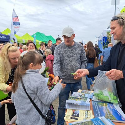 Ein Mann in schwarzem Shirt und mit schwarzem Sakko h&auml;lt in der linken Hand eine durchsichtige T&uuml;te mit Gummib&auml;rchen in gr&uuml;nen Plastikt&uuml;tchen. Ein T&uuml;tchen &uuml;berreicht er einem M&auml;dchen im grauen Pulli mit langen, dunkelblonden Haaren mit Pony, die zu einem Pferdeschwanz zusammengebunden sind.