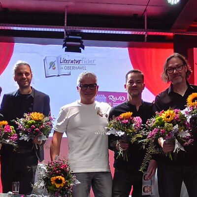 Die drei Autoren Richard Brandes, Sven Stricker und Andreas Winkelmann, der Moderator Vincent Kliesch und die drei Musiker der Pankeperlen auf der B&uuml;hne mit den zuvor &uuml;berreichten Blumenstr&auml;u&szlig;en.