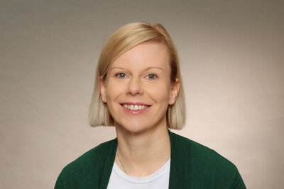 Eine Frau mittleren Alters mit blonden, kinnlangen Haaren, gr&uuml;nen Augen und gr&uuml;ner Jacke und wei&szlig;em Shirt. 