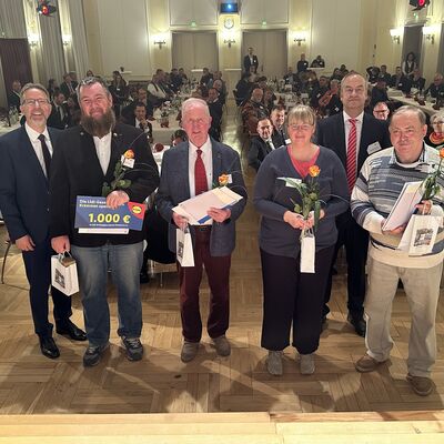 Im Vordergrund vier M&auml;nner und eine Frau. Im Hintergrund Menschen an Festtafeln in einem Festsaal.