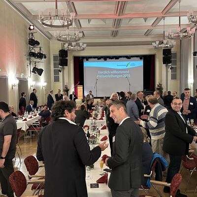 M&auml;nner und Frauen in einem feierlichen Festsaal. Im Hintergrund eine B&uuml;hne mit Leinwand.