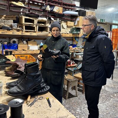 Ein Mann und eine Frau in Winterjacken stehen in einer Fabrikhalle hinter einem Produktionstisch aus Holz, auf dem Schuhe gefertigt werden.