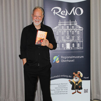 Der Autor Marc Raabe vor dem Roll-up Banner des ReMO - Regionalmuseum Oberhavel nach seiner Lesung am 24.11.2025 w&auml;hrend er seinen Thriller "Die Nacht" hochh&auml;lt.