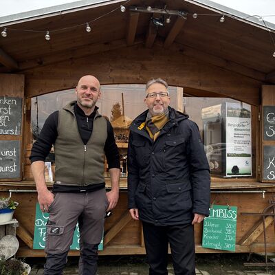 Zwei M&auml;nner stehen vor einen Holzh&uuml;tte mit Schildern.