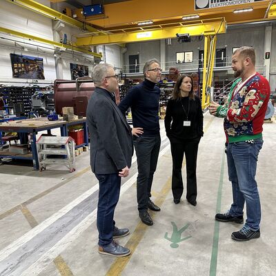 Drei M&auml;nner und eine Frau ganz in schwarz gekleidet stehen in einer Elektromotorenfabrikhalle im Halbkreis und unterhalten sich.