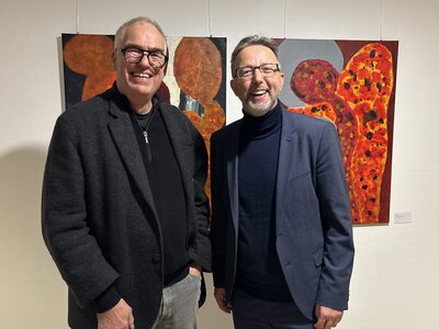 Zwei M&auml;nner mittleren Alters stehen vor einer wei&szlig;en Wand an der zwei farbenfrohe Bilder h&auml;ngen.