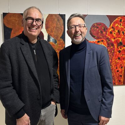 Zwei M&auml;nner mittleren Alters stehen vor einer wei&szlig;en Wand an der zwei farbenfrohe Bilder h&auml;ngen.