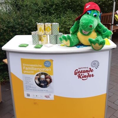 Ein gr&uuml;nes Pl&uuml;schkrokodil sitzt auf ein Messestand vom Netzwerk Gesunde Kinder.