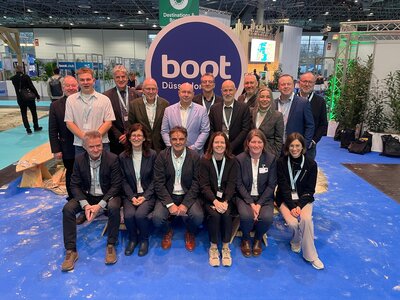 M&auml;nner und Frauen, dunkel gekleidet, sitzen in Zweierreihen vor einem Schild mit der Aufschrift boot D&uuml;sseldorf. 