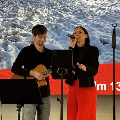 Ein Mann mit einer Gitarre musiziert auf einer B&uuml;hne neben einer dunkelhaarigen Frau mit Mikrofon in der Hand. Die Frau tr&auml;gt einen schwarzen Blazer und eine leuchtendrote, lange Hose.