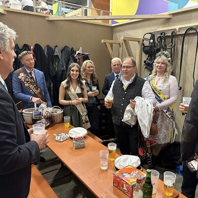 M&auml;nner und Frauen stehen um einen Holztisch auf einem Messestand und halten zum Teil Getr&auml;nke in den H&auml;nden.