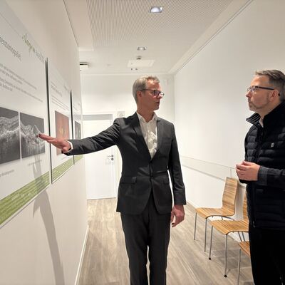 Zwei M&auml;nner mittleren Alters stehen im Flut einer modernen Wohnung oder Arztpraxis. An der Wand h&auml;ngen medizinische Bilder rund ums Thema Auge.