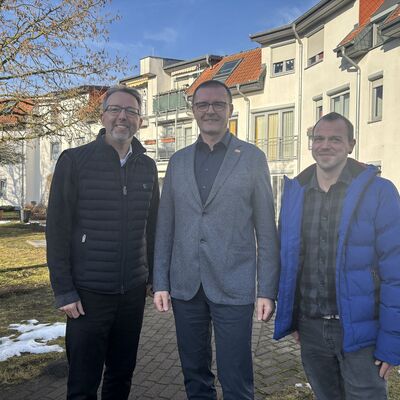 Drei M&auml;nner stehen vor einer cremefarbenen Geb&auml;udeanlage.