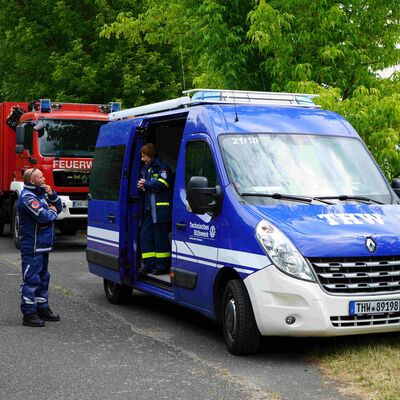 Ein blaues THW- und rotes Feuerwehrfahrzeug parken seitlich unter B&auml;umen an einer Landstra&szlig;e.
