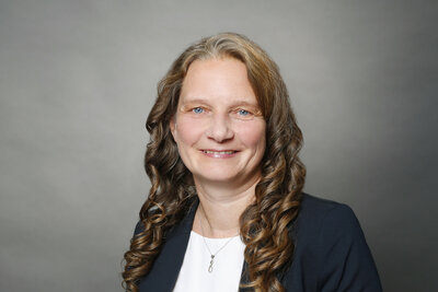 Eine Frau mittleren Alters mit langen dunkelblonden Locken, blauen Augen und dunkelblauer Jacke und wei�em Oberteil.