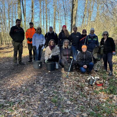 M&auml;nner und Frauen stehen in und hocken in Zweierreihen in einem Waldgebiet. Sie haben Eimer und M&uuml;llsammelzangen dabei.